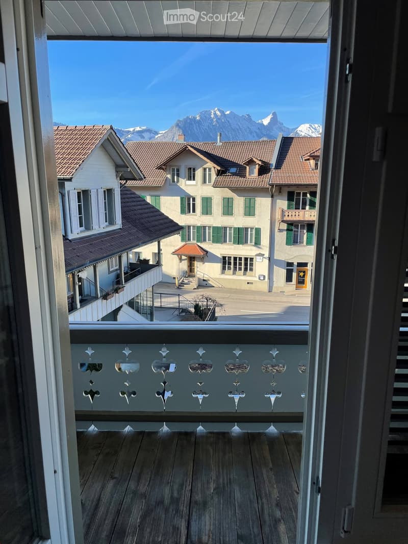 Balcony with wooden decking, ornate metal balustrade, overlooking a town square with traditional houses and snow-capped mountains in the background.