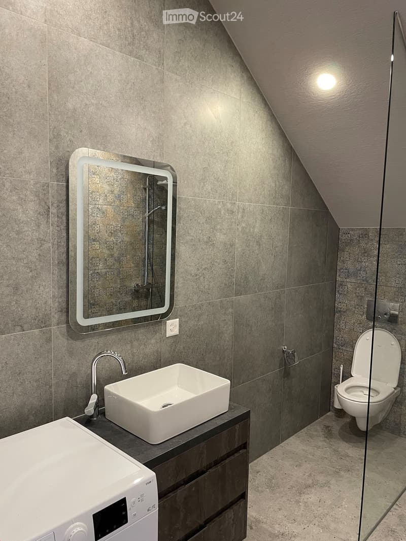 Modern bathroom with a white sink, chrome faucet, illuminated mirror, toilet, and a washing machine. Grey tiled walls and a sloped ceiling.
