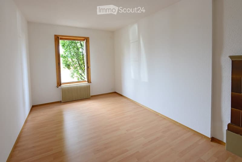Empty room with wooden flooring, white walls, a window with wooden frame, radiator, and a small fireplace on the right side.