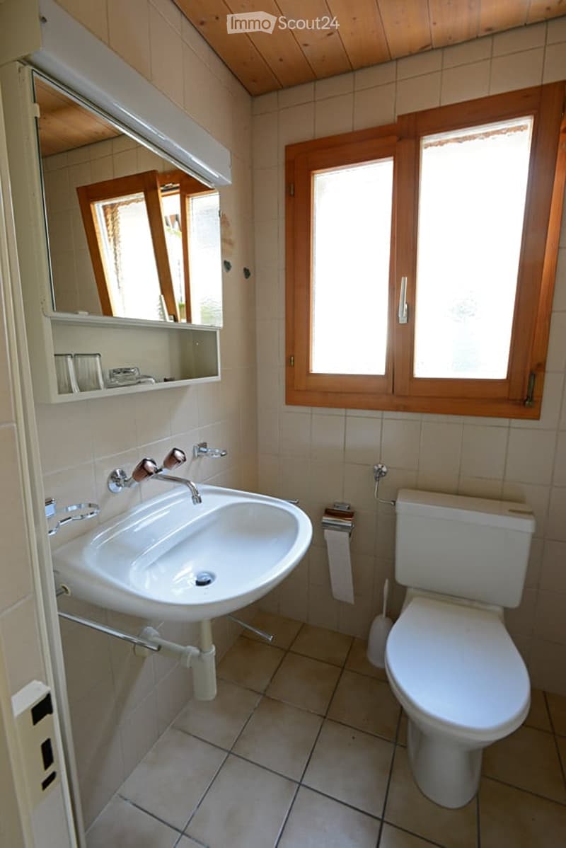 Bathroom with a white sink, mirror, toilet, and window with wooden frame. The room features tiled walls and floor, and a small shelf above the sink.