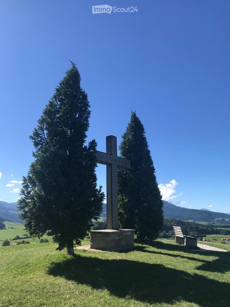 A serene outdoor setting featuring a large cross monument on a stone base, surrounded by two tall, well-maintained evergreen trees. A wooden bench is positioned to the right, with a scenic mountain backdrop and open green fields extending into the distanc