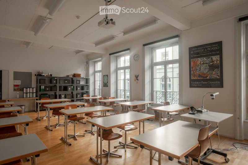 Classroom with wooden floors, large windows, multiple tables and chairs, shelves with storage boxes, a projector, and a clock on the wall.