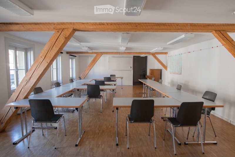 Bright, spacious room with wooden floors and exposed wooden beams. Multiple tables and chairs are arranged in rows, suitable for a classroom or meeting space. Large windows allow natural light to fill the room.