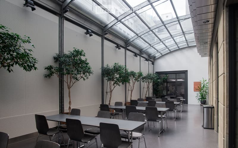 Modern indoor space with large glass ceiling and walls, featuring multiple tables and chairs arranged for seating, several potted trees, and a trash can near the wall.