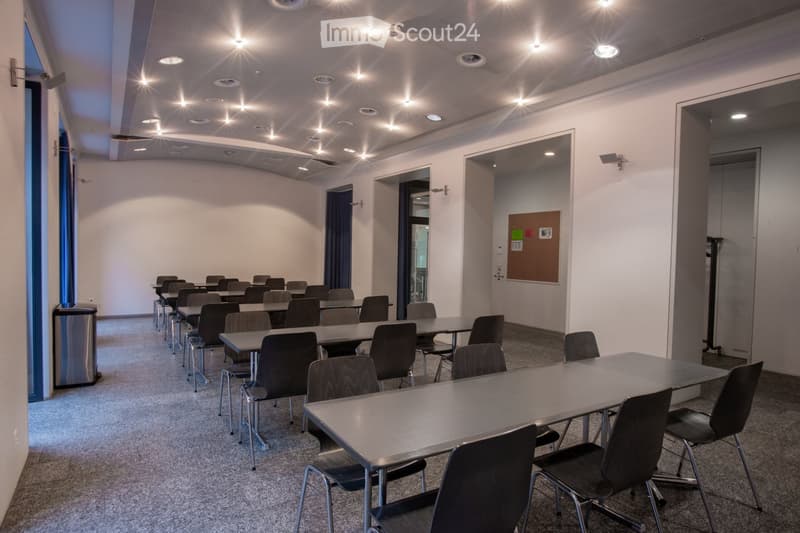 Modern, well-lit room with multiple tables and chairs arranged in rows, a trash can, and a bulletin board on the wall.