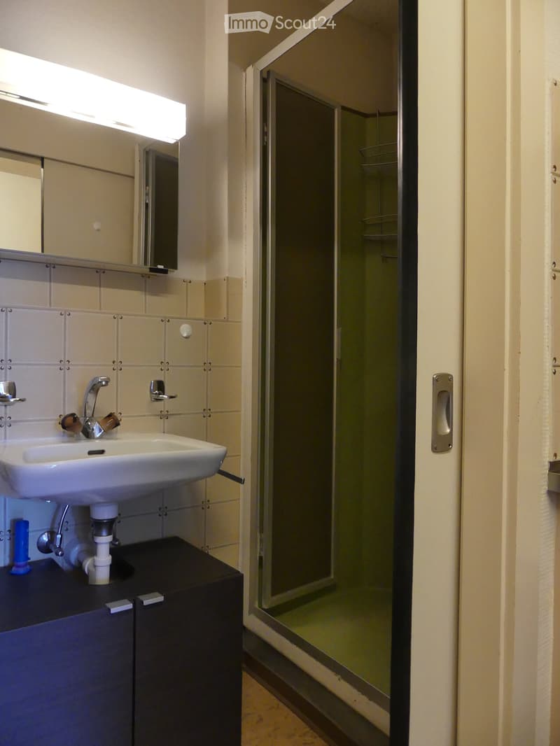 Bathroom with a white sink, mirror, and a glass-enclosed shower. The sink has a modern faucet and is mounted on a dark-colored cabinet. The walls are tiled in light beige, and the floor is tiled in a light color.
