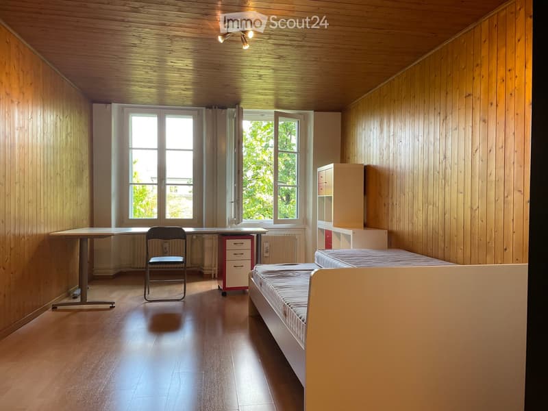 A small, furnished room with wooden walls and ceiling, featuring a single bed, a desk with a chair, a small cabinet, and a drawer unit.
