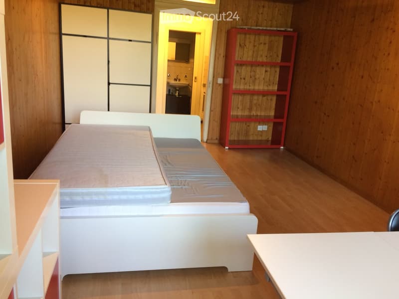 A furnished room with a bed, a table, and a shelf. The room has wooden flooring and a partially visible bathroom in the background.