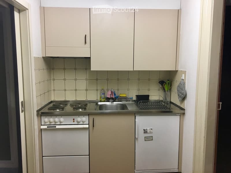 Compact kitchen with white cabinets, a four-burner stove, a sink, and a small refrigerator. The walls are tiled, and various kitchen utensils and cleaning supplies are visible on the counter.