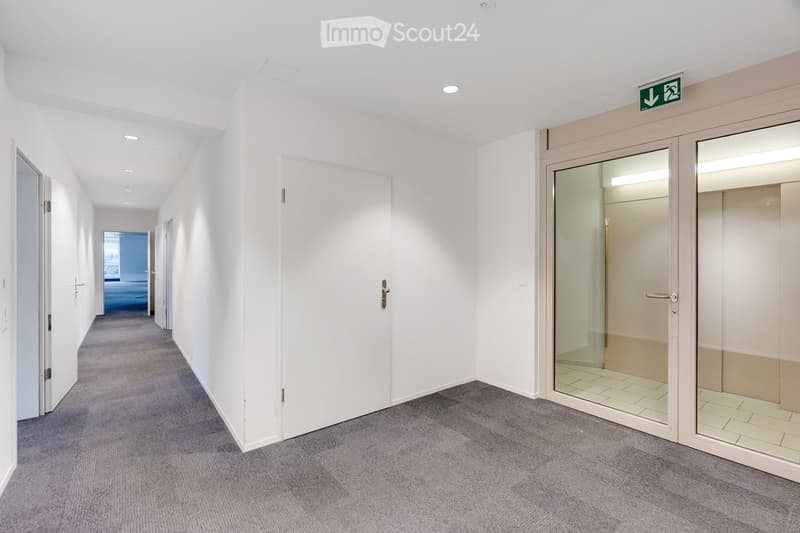 Clean, modern hallway with white walls, grey carpet, multiple doors, and a visible emergency exit sign.