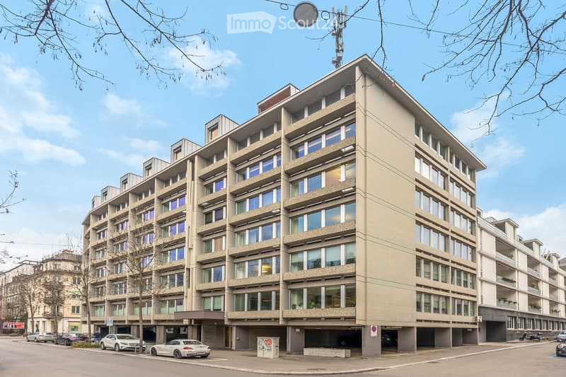 Multi-story residential building with large windows and balconies, located on a street with parked cars and trees.