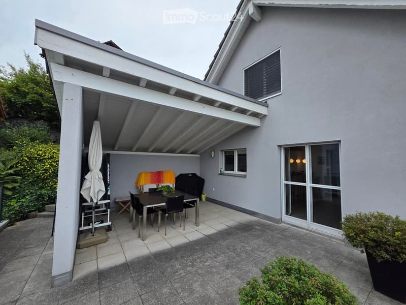 Covered patio area with a dining table, chairs, umbrella, bench, and potted plant, adjacent to a modern house with large windows and a view of greenery.