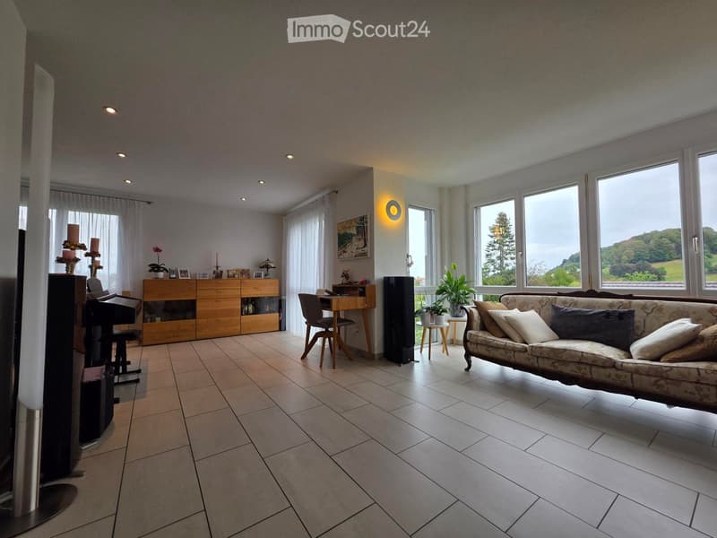 Bright living room with large windows, a patterned sofa, a wooden cabinet, a small table with a plant, a piano, and a desk area with a chair.