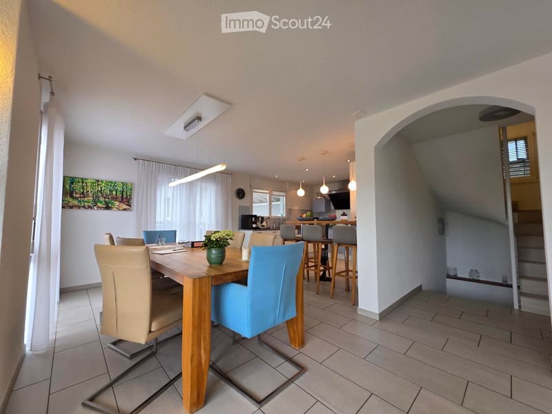 Bright dining area with a wooden dining table surrounded by chairs and stools, a flower vase in the center, and a modern kitchen area visible in the background.