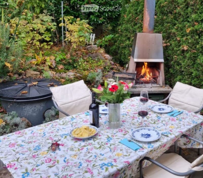 Garden area with a dining table set for two, surrounded by chairs, a vase with flowers, and a lit wood stove in the background.