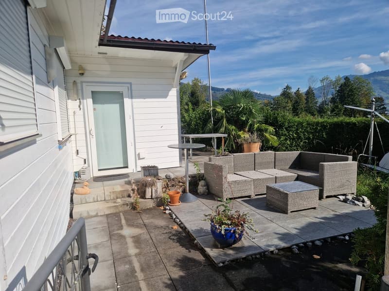 Outdoor patio with wicker furniture, small table, potted plants, and a view of the surrounding greenery and mountains.