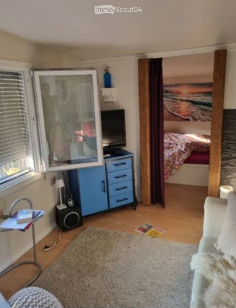 A cozy living room with a sofa, a small table, a cabinet with a TV, a lamp, a speaker, a mirror, and a bed visible in the background.