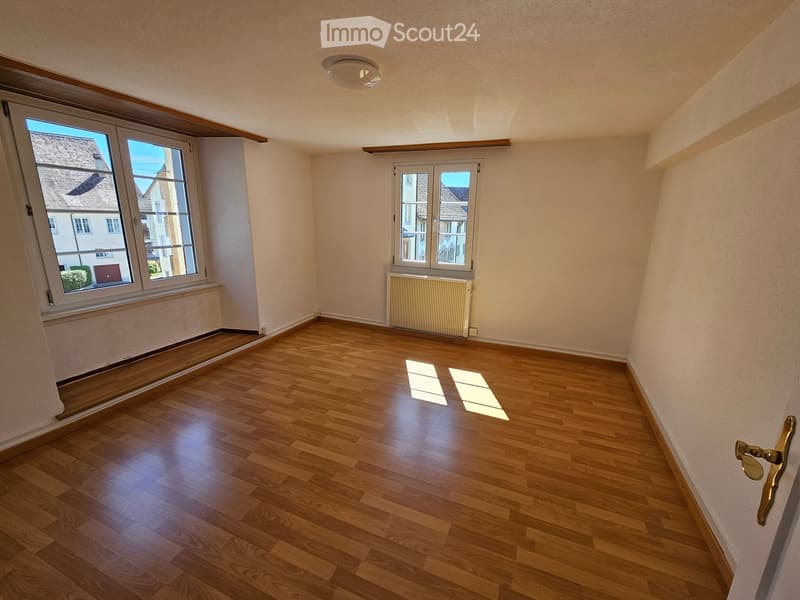 Empty room with wooden flooring, large windows, and a radiator. No furniture or equipment present.
