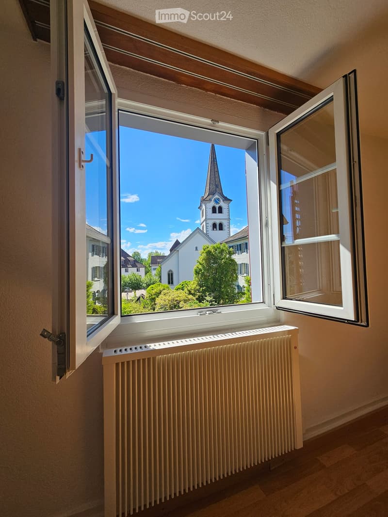 Empty room with wooden floor, beige walls, large window with open shutters, radiator below the window, view of a church tower and surrounding buildings.