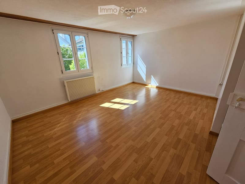 Empty room with wooden flooring, white walls, and two windows allowing natural light. A radiator is visible on one wall.