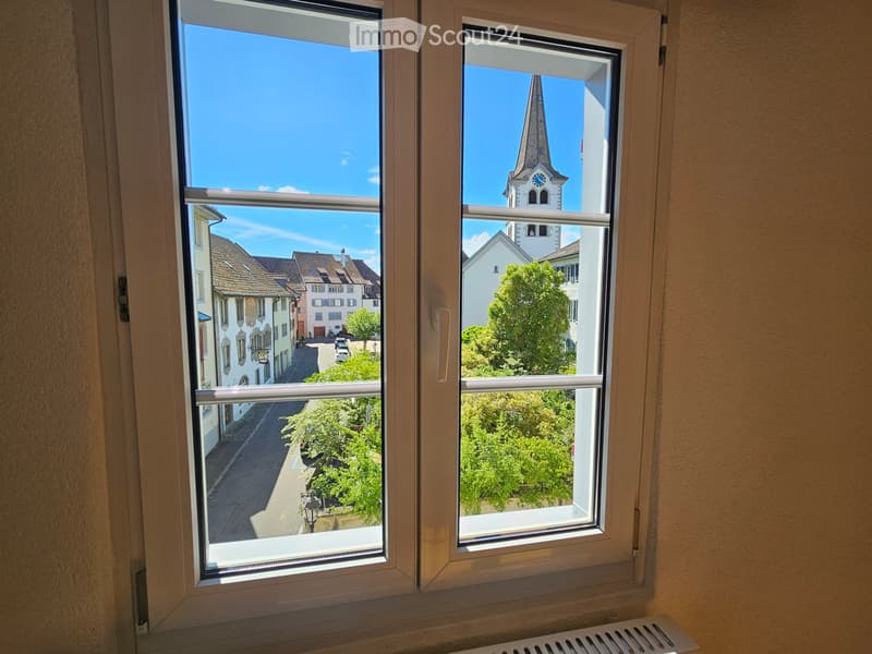 View from a window showing a town street with buildings and a church tower in the background.