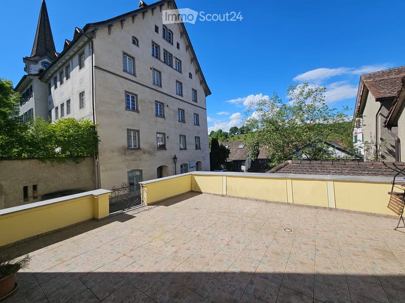 Balcony with stone tiles, yellow walls, metal railing, and a view of a historic building with a spire.