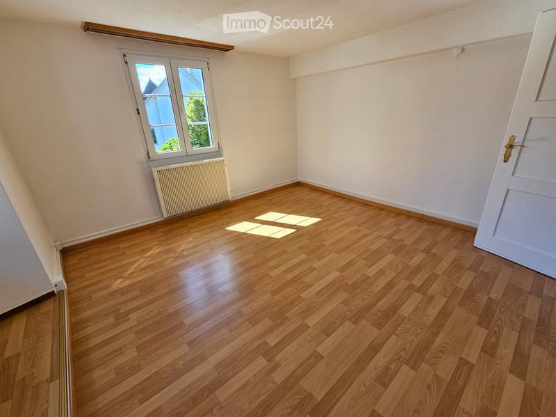 Empty room with wooden flooring, white walls, and a window with a radiator below it. The room has a slanted ceiling typical of an attic space.