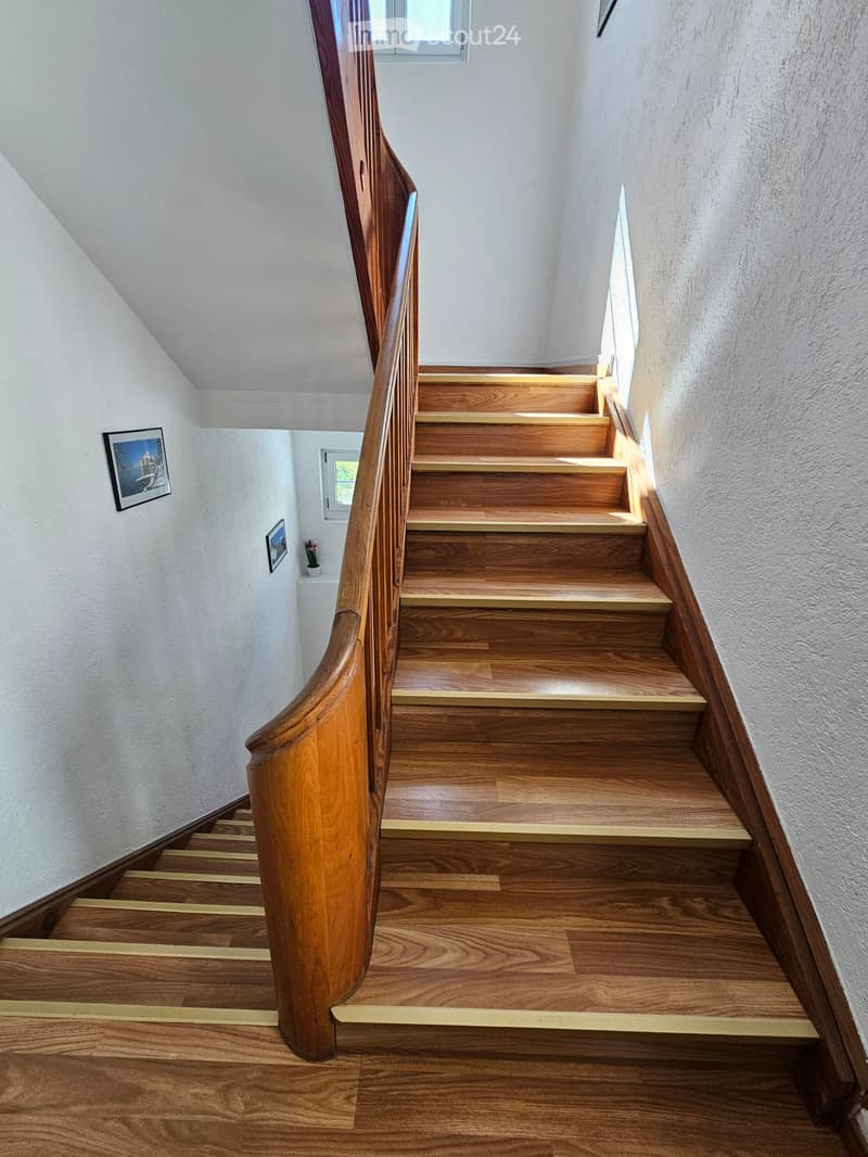 Spiral wooden staircase with polished wooden steps and handrail, white walls, small windows, and framed pictures on the wall.