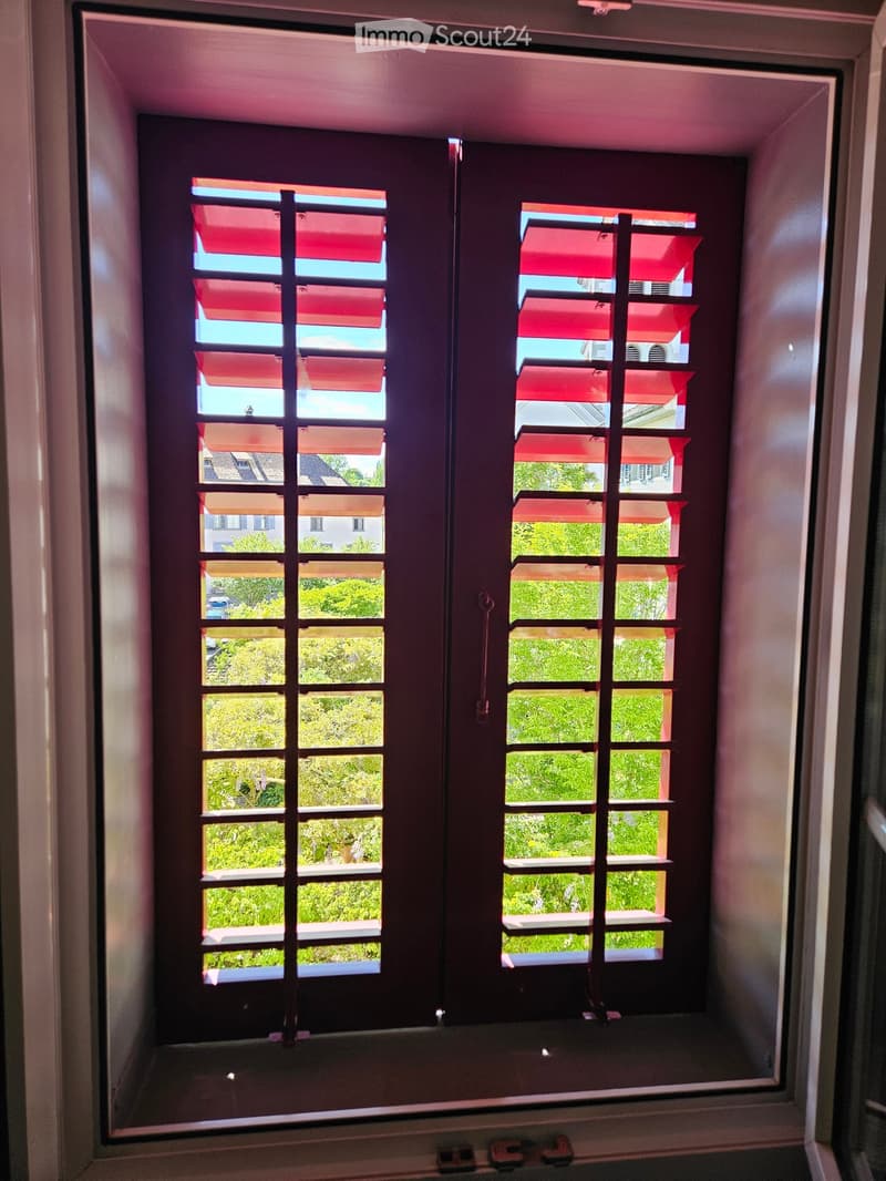 An unfurnished room with a window featuring red shutters, offering a view of greenery and neighboring buildings.