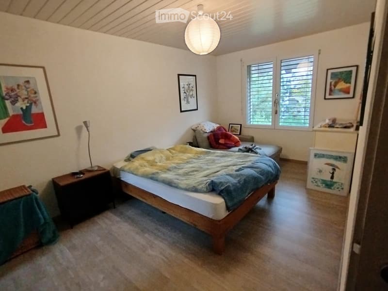 Cozy bedroom with a wooden bed, nightstand, lamp, and a small seating area with a chair and additional bedding.