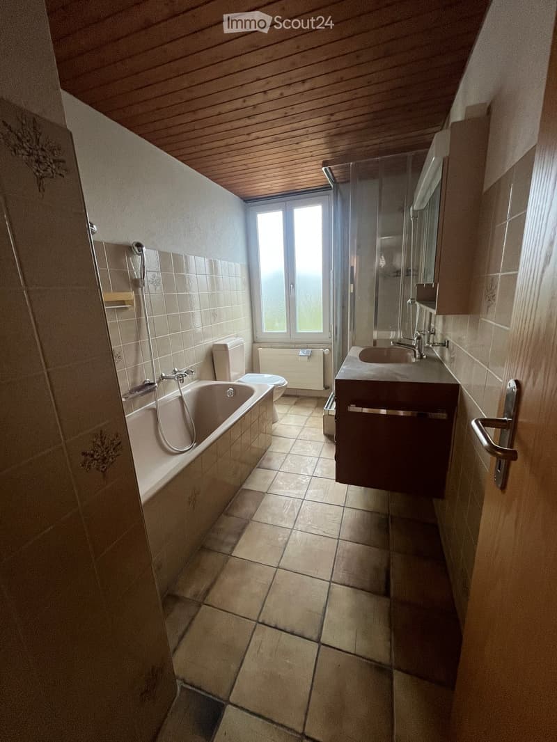 Bathroom with a bathtub, toilet, sink, and shower area. The room features tiled walls and floor, a window, and a wooden ceiling.