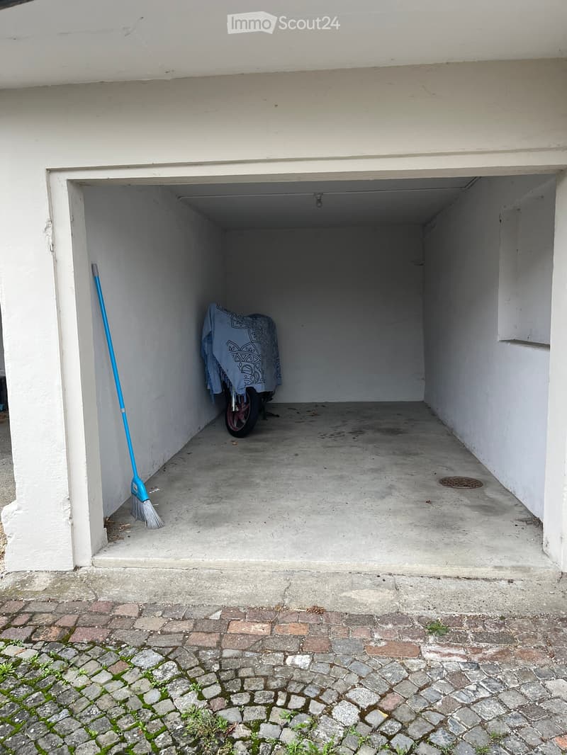 Garage with white walls, concrete floor, broom leaning against wall, covered object in the center.