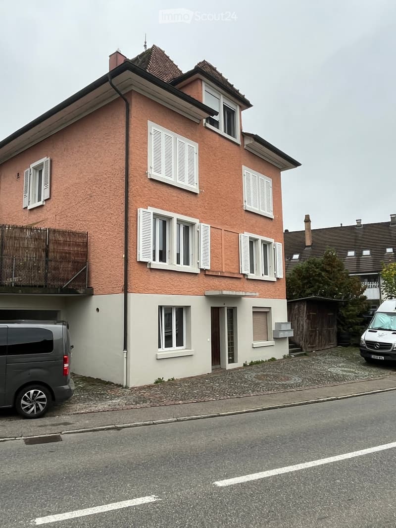 Two-story house with pink facade, white shutters, and multiple windows. Features a garage and a small balcony on the left side.