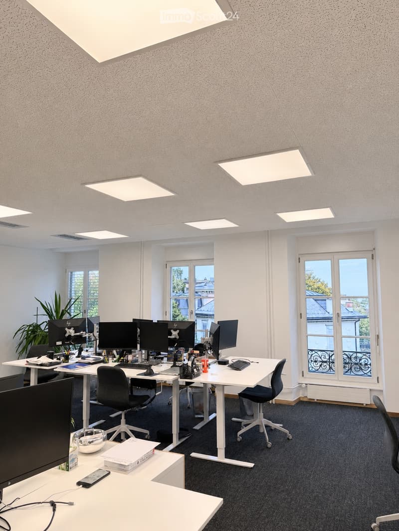 Modern office space with multiple desks, computer monitors, chairs, and office supplies. Large windows provide natural light, and the room features contemporary furniture and decor.
