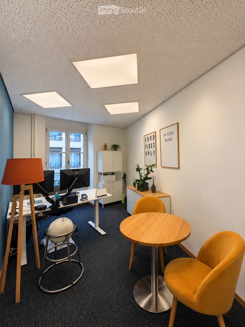 Modern office space with a standing desk, ergonomic chair, round meeting table with two yellow chairs, a floor lamp, a small cabinet, and decorative plants.