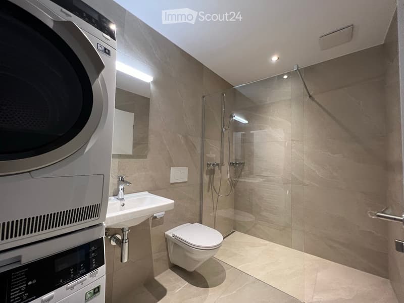 Modern bathroom with a sink, toilet, shower, and washing machine. Features include beige tiles, a glass shower enclosure, and built-in lighting.