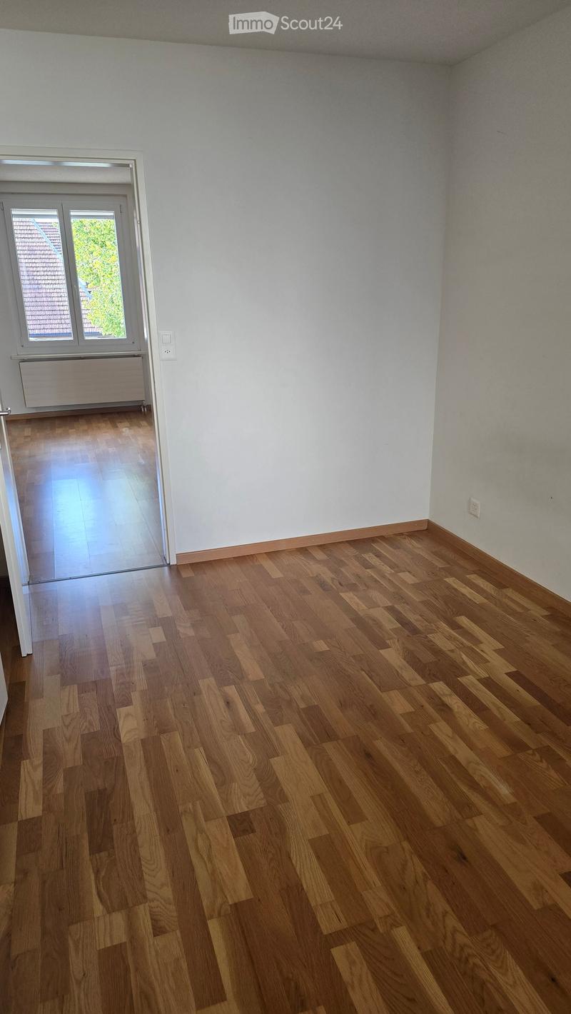 Empty room with wooden flooring, white walls, and a large window with a view of greenery outside. The room has a radiator and a door leading to another space.