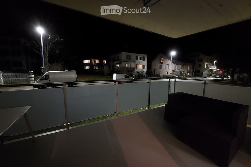 Balcony with glass railing, small table, and view of street with parked vans and residential buildings.