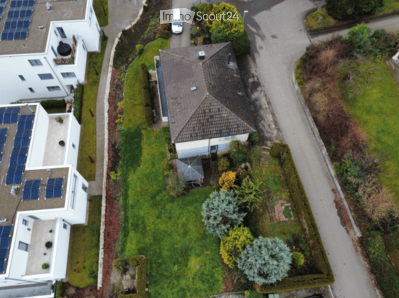 Aerial view of a residential area showing a single-family house with a tiled roof, surrounded by greenery and neighboring houses with solar panels.