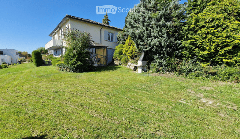 Well-maintained garden with a large, single-story house in the background. The house features a balcony, multiple windows, and a small shed. The garden is lush with various trees and shrubs, and a stone bench is visible near the house.