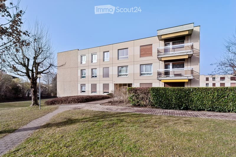 Modern multi-story residential building with beige facade, multiple windows, and balconies. The building is surrounded by a well-maintained lawn, hedges, and a paved pathway.