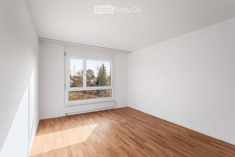 Empty room with white walls, wooden flooring, and a large window with white frame and radiator below it.