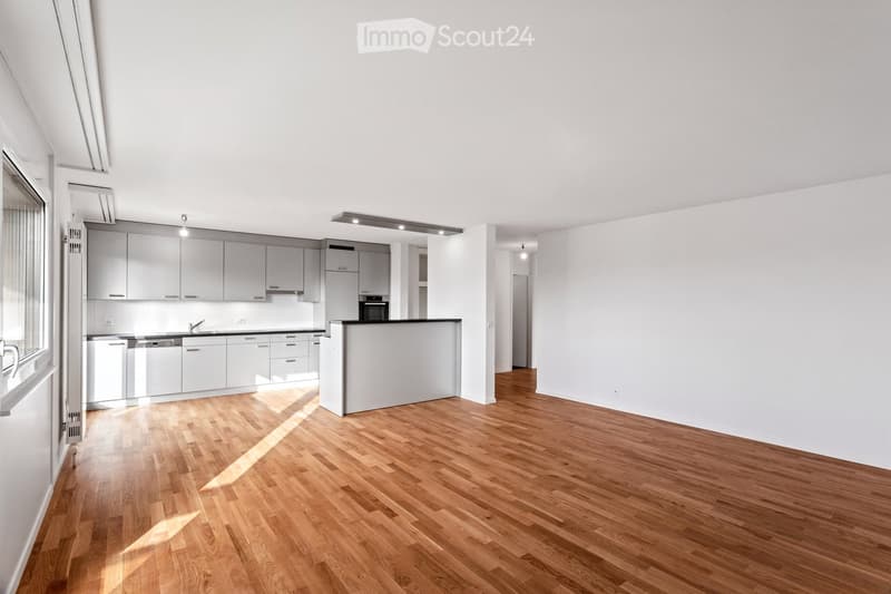 Empty room with light wooden flooring, white walls, and a modern kitchen area featuring grey cabinets, a sink, and built-in appliances.