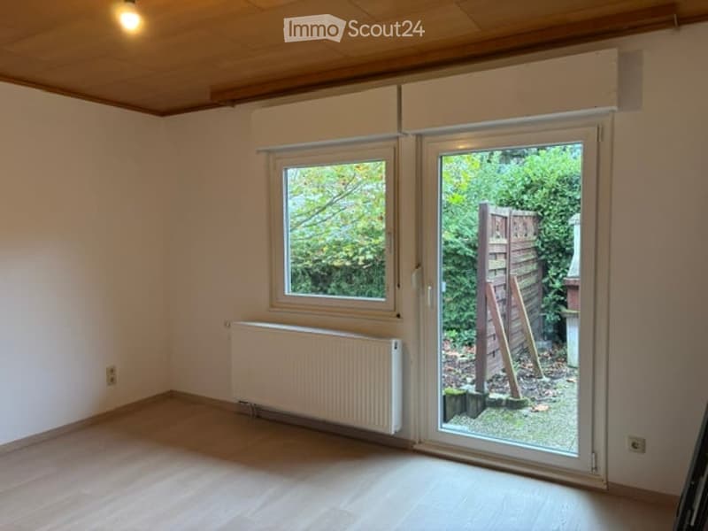 Empty room with wooden flooring, white walls, large window, sliding glass door leading to outdoor area, radiator, and visible ceiling light fixture.