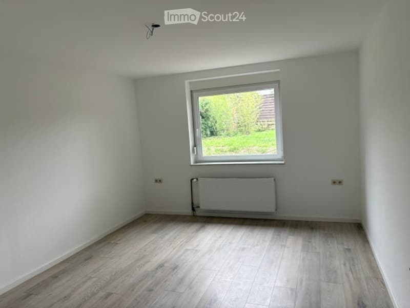 Empty room with light grey walls, wooden flooring, a single window with white frame, and a radiator below the window.