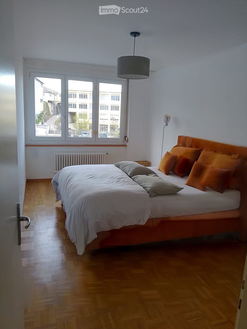 Bright bedroom with wooden flooring, large window offering a view of the street, a neatly made bed with pillows, a radiator below the window, and wall-mounted and pendant lamps for lighting.