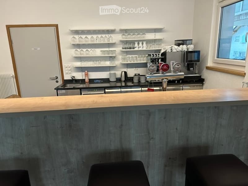 Modern bar area with wooden counter, shelves holding glasses, coffee machine, and stools.
