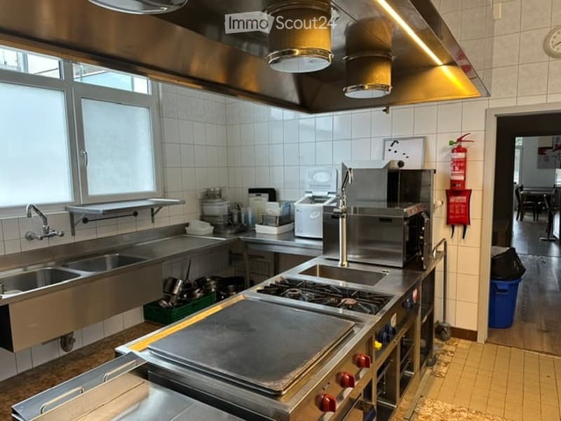 Commercial kitchen with stainless steel appliances, including a sink, microwave, oven, stove, and refrigerator. The space features large windows, tiled walls, and a well-organized counter area.