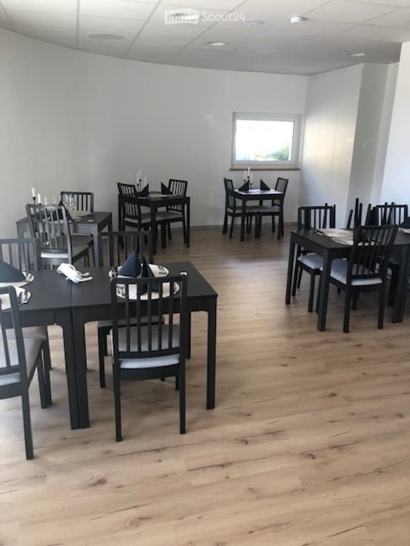 A modern, minimalist restaurant or dining area with wooden floors, black tables, and matching chairs arranged neatly. Each table is set with napkins, candles, and glasses, creating a clean and inviting atmosphere.