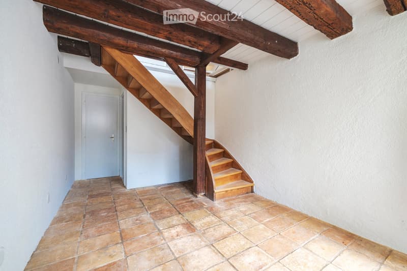 A narrow hallway with exposed wooden beams and a wooden staircase leading upwards, featuring tiled flooring and white walls.